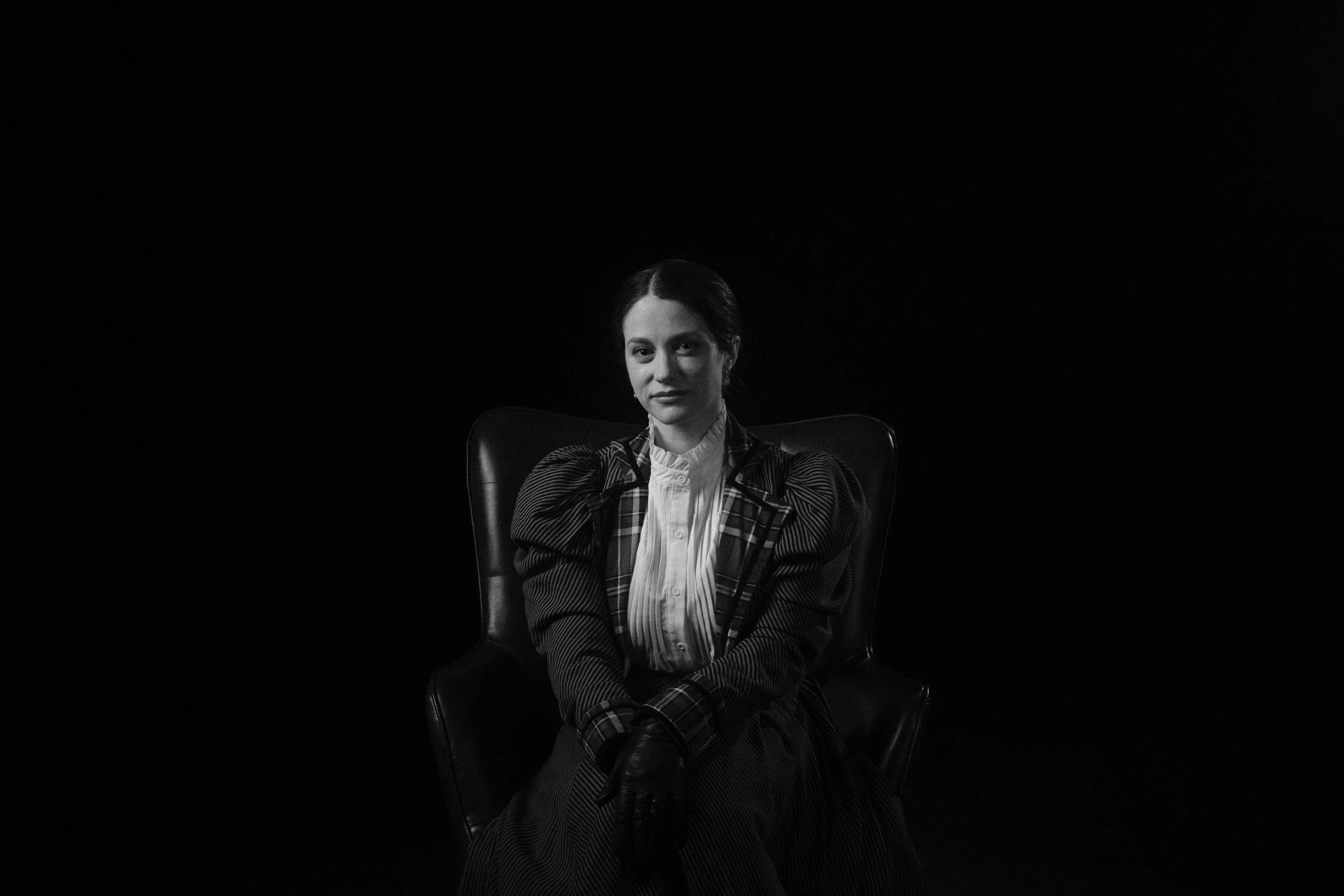 Black-and-white portrait of a woman in period costume seated in a chair against a dark background.